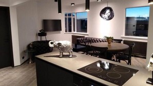 A modern kitchen with a central island, pendant lights, a dining area with a round table, and a large window.
