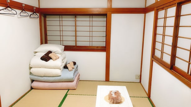 A traditional Japanese room with tatami flooring, a futon bed, a small table with a decorative item, and a shoji screen window. - Guanjing Apartment (Tokyo)