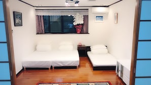 A small, well-lit room with two single beds, a nightstand, a window with curtains, and a colorful rug on the wooden floor. - Guanjing Apartment (Tokyo)