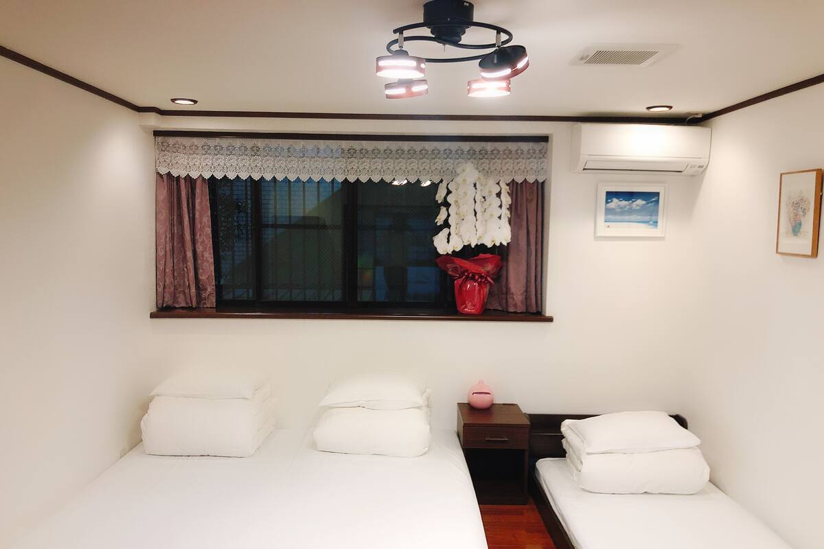 A bedroom with two beds, a window with a lace curtain, a ceiling fan, and a framed picture on the wall.