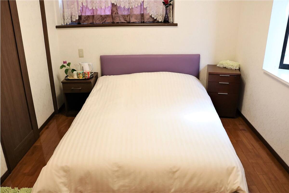 A neatly made bed with a purple headboard, a bedside table with a tray of items, and a small drawer unit.