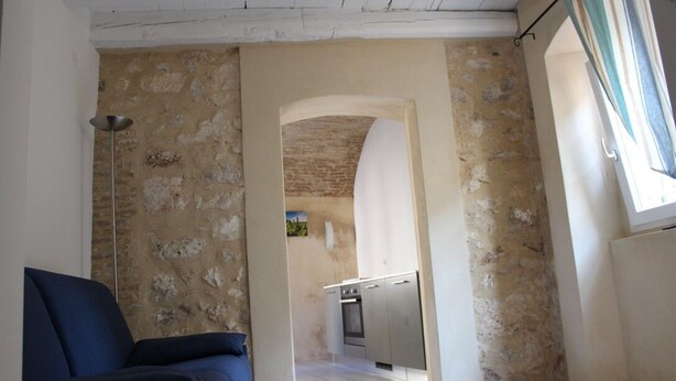 Una habitación con un sillón azul, una cocina al fondo y una ventana con cortinas.