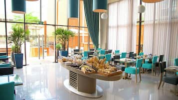 Montaje de bufé con diferentes alimentos, rodeado de mesas y sillas en un área de comedor luminosa y moderna con ventanas grandes y plantas de interior.