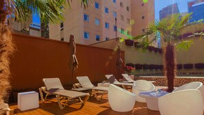 Un salon sur le toit, avec des fauteuils blancs et une petite table, entouré de plantes en pot et offrant une vue sur la piscine.