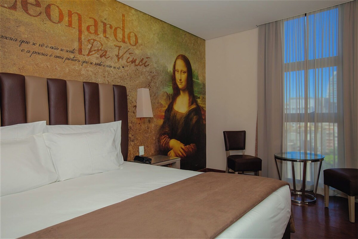 A hotel room with a bed, a chair, a small table, and a large painting of the Mona Lisa on the wall.