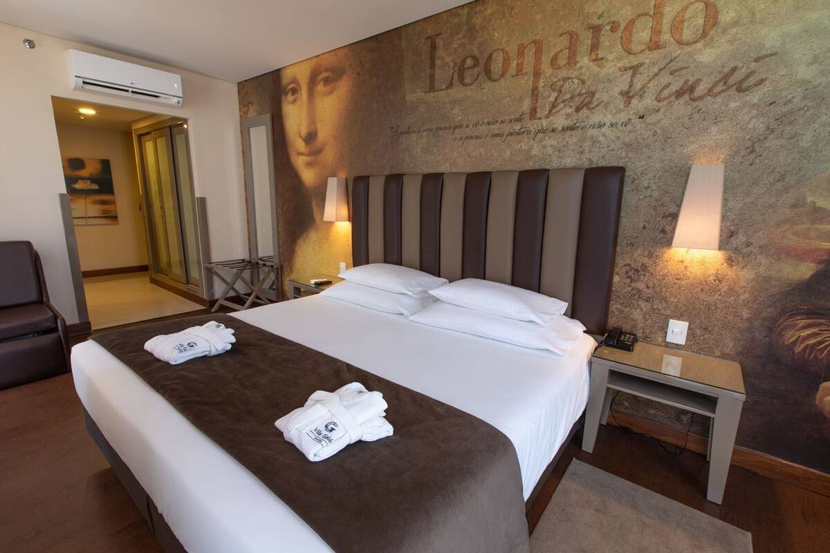 A hotel room with a large bed, two bedside tables, a desk, and a chair. There is a painting on the wall and an air conditioning unit on the ceiling.