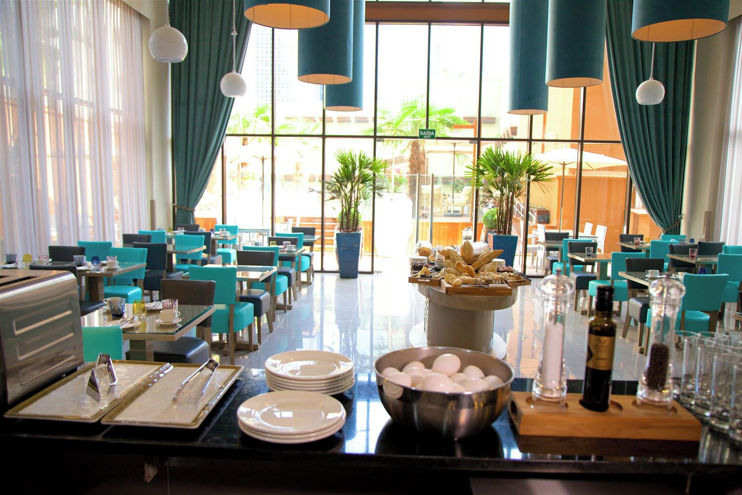 A restaurant with teal chairs and tables, a buffet with pastries, and a view of palm trees through large windows.