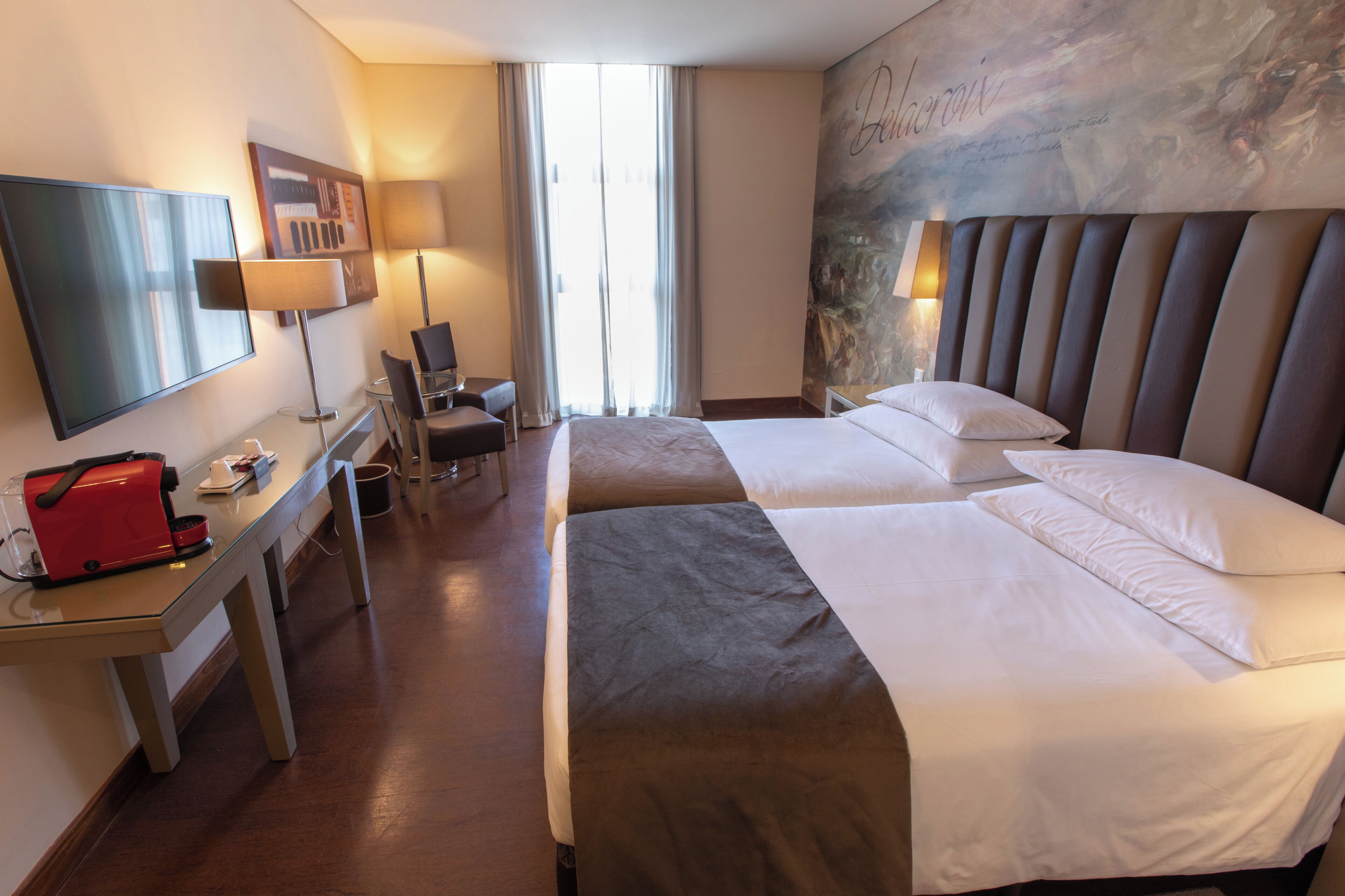 Une chambre d’hôtel avec deux lits, un bureau sur lequel se trouve un sac rouge, une télévision et un mur orné d’une fresque.