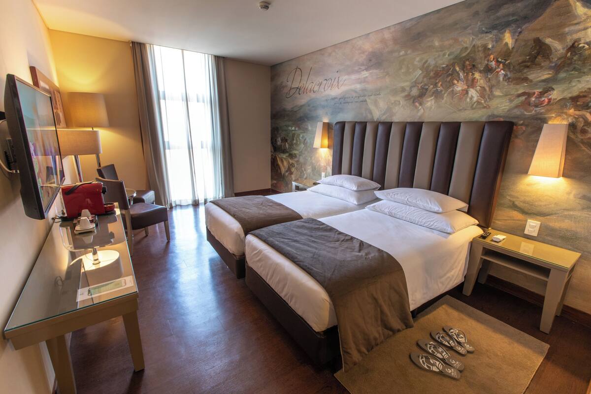 A hotel room with two beds, a desk with a lamp, a TV, and a painting on the wall.