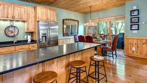 A kitchen with wooden cabinets, a stainless steel refrigerator, a large island with stools, and a dining area with chairs and a chandelier.