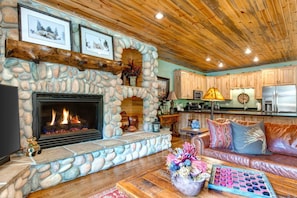 A stone fireplace with a fire, wooden ceiling, kitchen area, and a leather sofa.