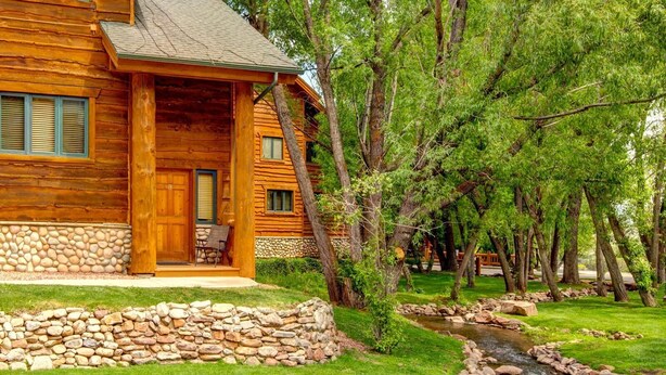 A log cabin with a stone foundation, a porch with a chair, and a stream running through the property.