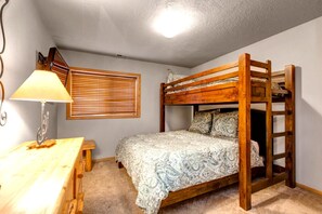 A bunk bed room with a wooden desk and lamp, a window with blinds, and a ceiling light.