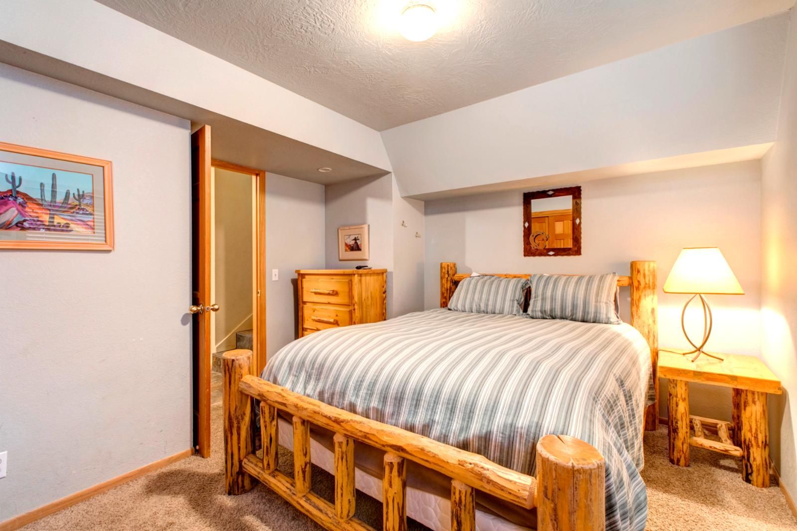 A bedroom with a wooden bed frame, a bedside table with a lamp, a dresser, and a door leading to another room.