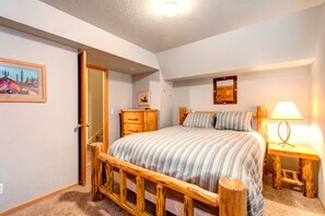 A bedroom with a wooden bed frame, a bedside table with a lamp, a dresser, and a door leading to another room. - Walk to Ski Slopes + Trails! Spacious 4-Bedroom Private Home with Fantastic Views, Hot Tub, A/C, and Wood Fireplace | PLA-1155 by KBM Resorts (Park City)