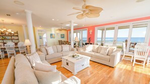 A spacious living room with a large sectional sofa, a coffee table, and a dining area with chairs. There is a balcony with a view of the ocean. - Private Pool, Elevator, Sleeps 20, Bob Sikes Cut! (St. George Island)