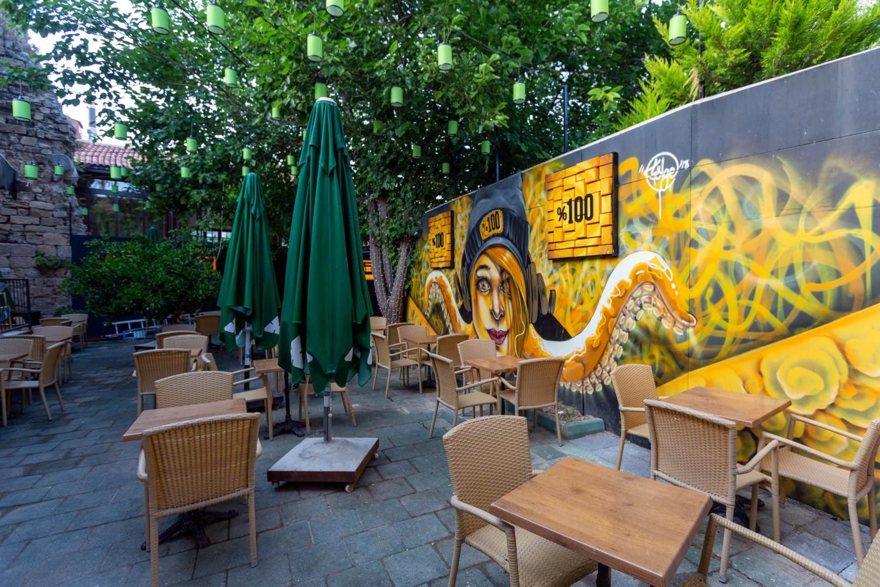 An outdoor seating area with wicker furniture and green umbrellas, a mural with a face and text, and a brick wall.