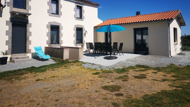 Un edificio de dos pisos con techo de tejas, un patio con mesa y sillas, y una sombrilla azul.