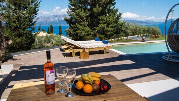 Een houten tafel met een fles wijn, glazen en een schaal fruit, gelegen bij een zwembad met uitzicht op bergen en zee.