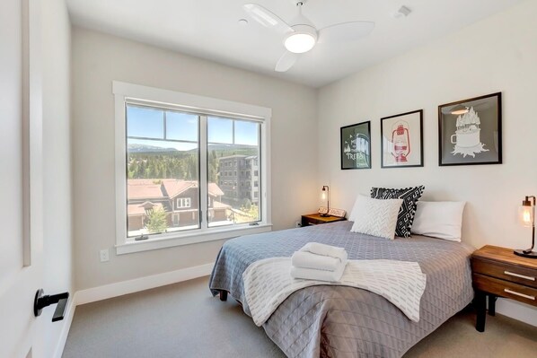 A bedroom with a large window, a bed with a quilt, a nightstand with a lamp, and three framed pictures on the wall.