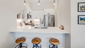 Une cuisine moderne avec des meubles blancs, des appareils en acier inoxydable et des luminaires suspendus. On y trouve trois tabourets en bois au comptoir.