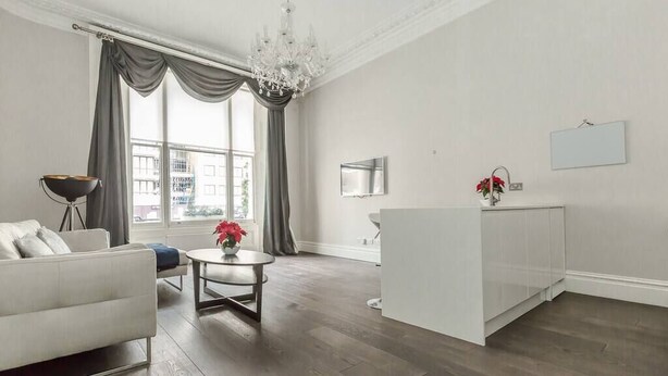 A modern living room with a white sofa, a round coffee table, a chandelier, and a large window with curtains. - Modern 1BR Period Property in Kensington (London)