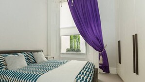 A bedroom with a bed, a window with a view of greenery, and a purple curtain. - Modern 1BR Period Property in Kensington (London)