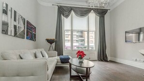 A modern living room with a white sofa, a round coffee table with a red flower, and a large window with curtains. - Modern 1BR Period Property in Kensington (London)