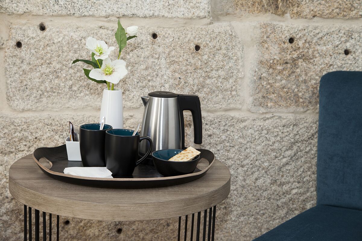 Una mesa redonda de madera con una bandeja que sostiene un jarrón blanco con flores, tazas negras, un hervidor plateado y un bol con pan plano.