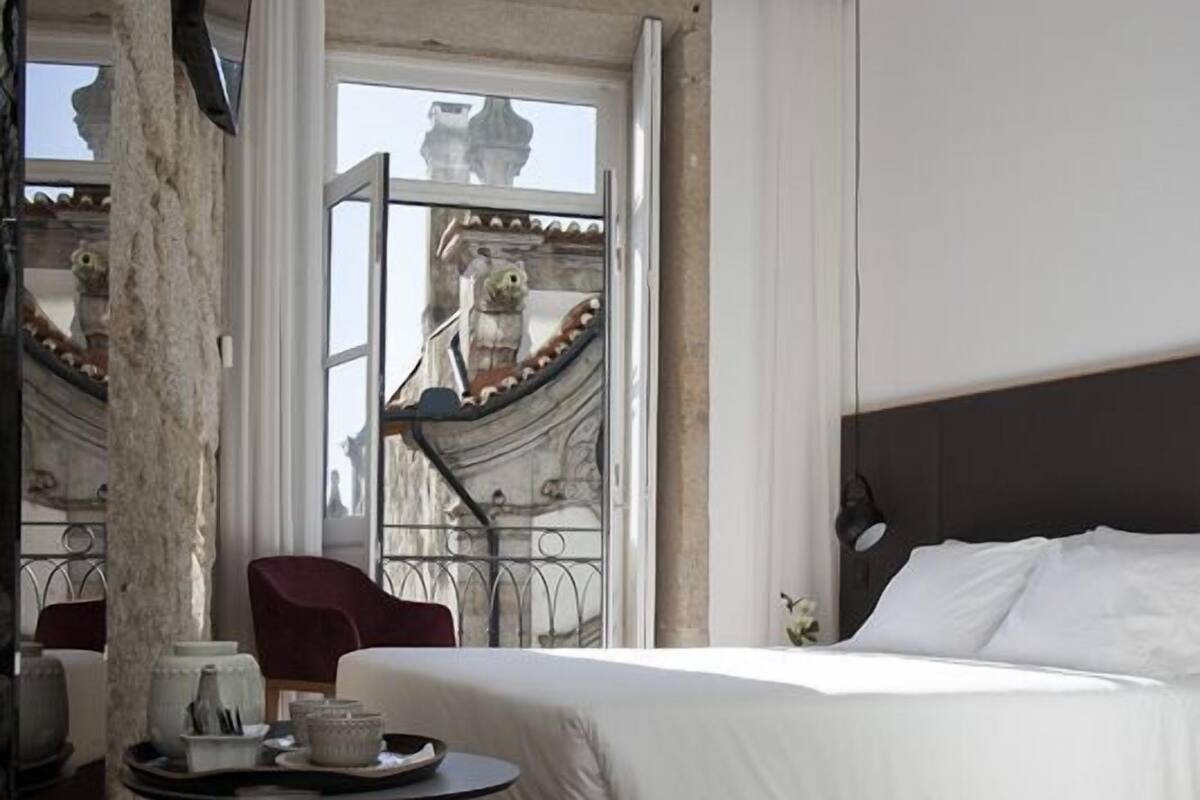 Una habitación de hotel moderna con una cama grande, una mesa redonda con una silla roja, y vistas a un edificio histórico a través de un ventanal amplio.