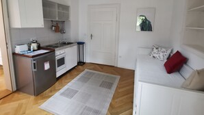 Eine kompakte Küche mit Kühlschrank, Herd und Spüle. Ein Esstisch mit Platzdeckchen und ein Sofa mit rotem Kissen sind vorhanden.