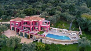 A multi-story pink villa with a swimming pool and outdoor seating area surrounded by lush greenery.