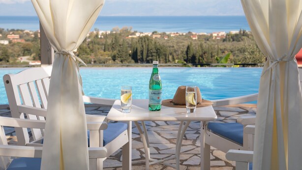 A terrace with a table set for two, overlooking a pool and a scenic view of the sea and hills.