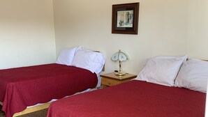 Habitación de hotel con dos camas, cada una con colcha roja, una mesita de noche con lámpara, ventilador de techo y un cuadro enmarcado en la pared.
