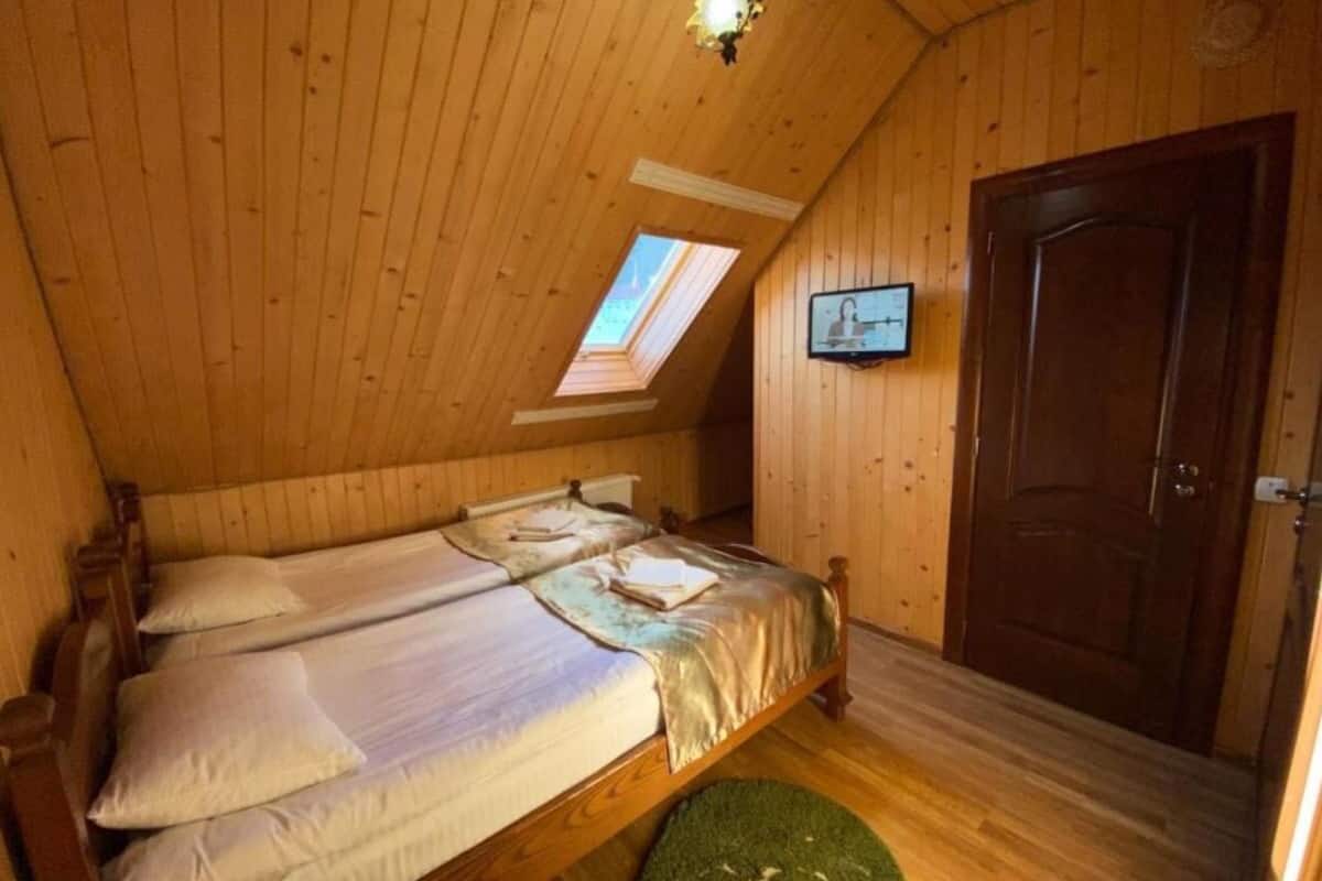 A cozy attic bedroom with wooden walls and a sloped ceiling. There are three single beds arranged in a row, a small skylight, and a door to the right.