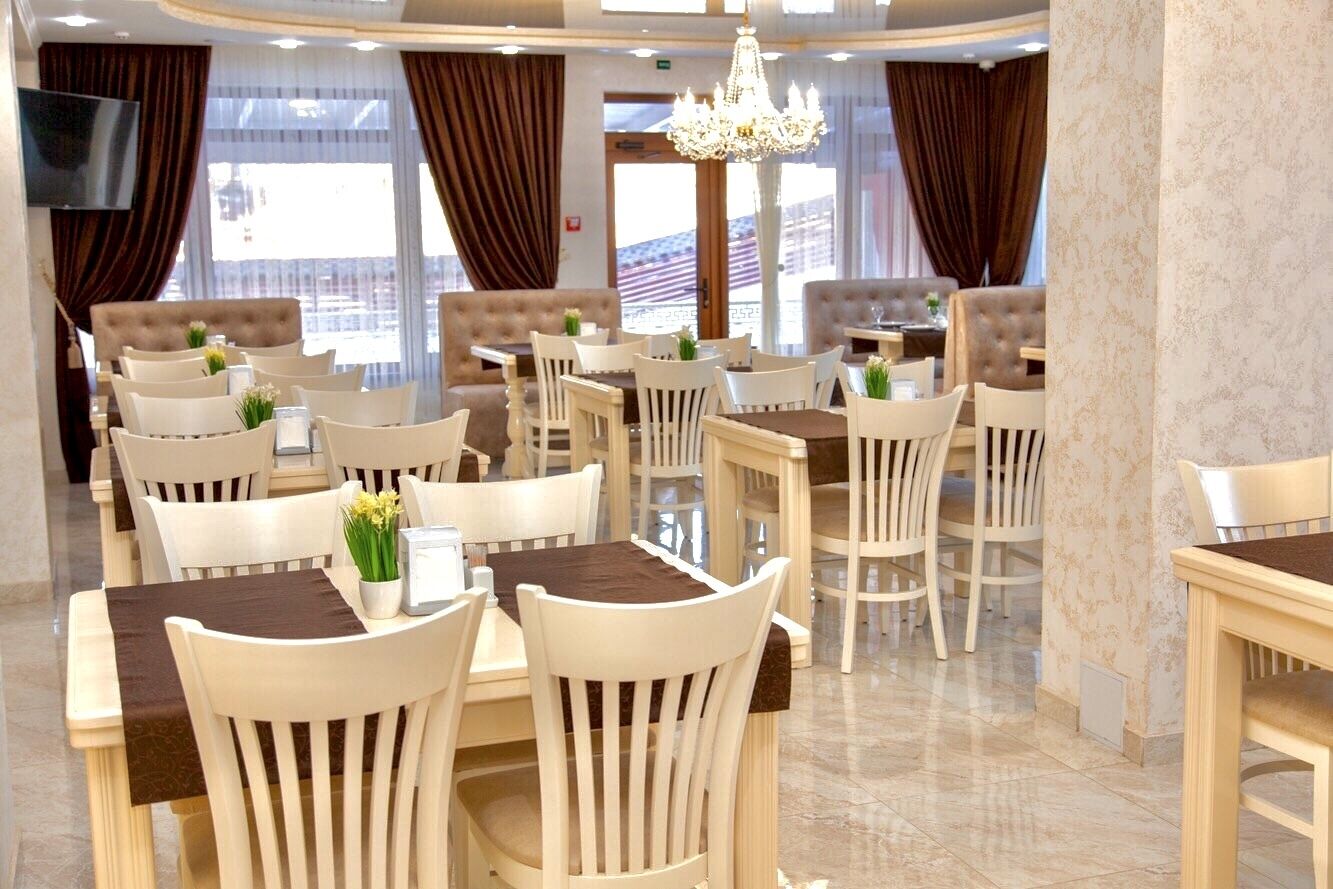 A hotel dining area with multiple tables and chairs, a chandelier, and large windows with curtains.