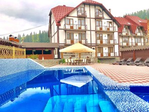 A hotel with a swimming pool, outdoor seating, and a mountain backdrop. - Villa Nikoletta (Bukovel)