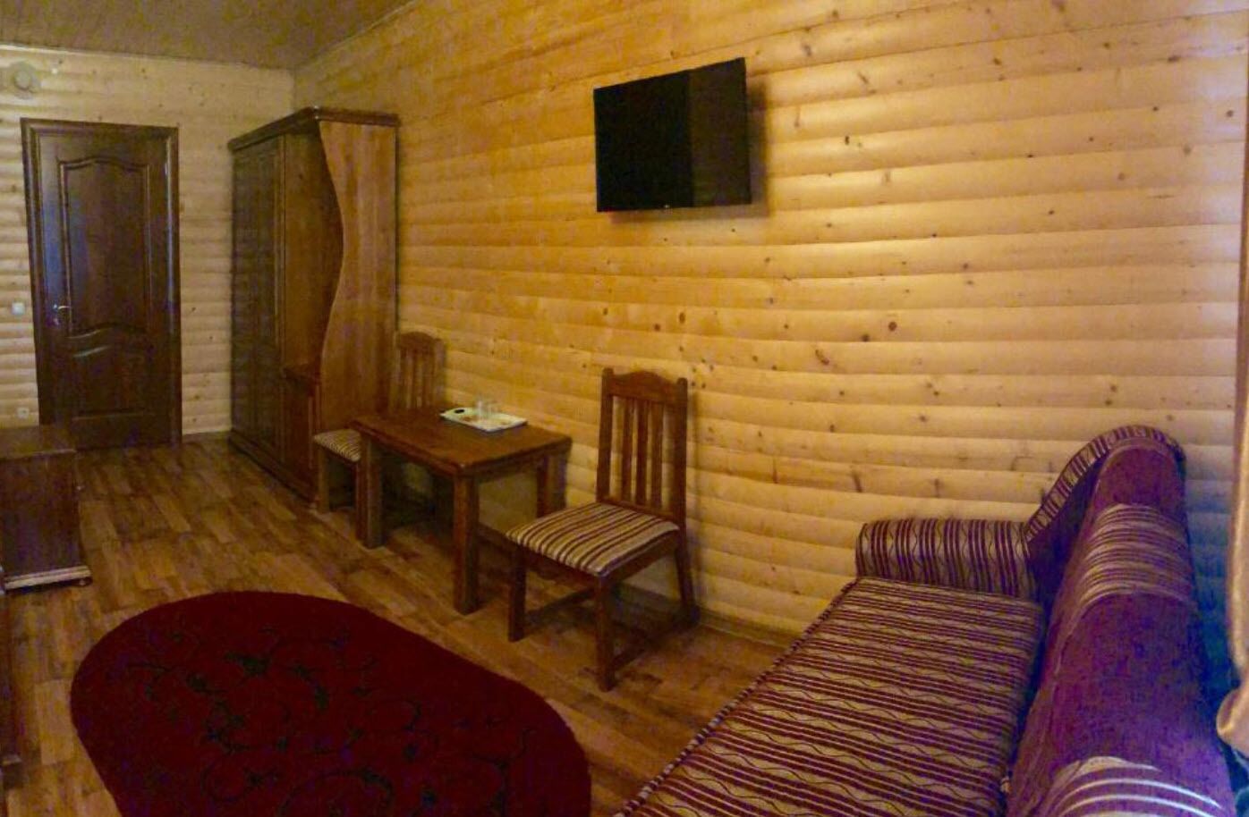 A log cabin interior with a wooden floor, a dining table with chairs, a sofa, and a television mounted on the wall.