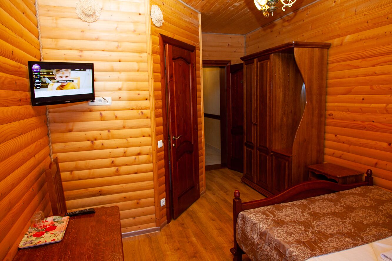 A wooden cabin room with a bed, a wooden wardrobe, a television mounted on the wall, and a small table with a remote control and a glass.