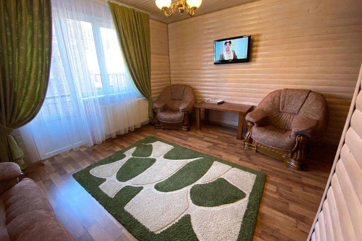 A room with wooden flooring, two leather armchairs, a green and white rug, a wooden bench, and a television on the wall.