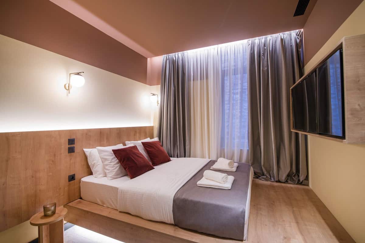 A modern hotel room with a wooden headboard, a large bed with white and red pillows, a bedside table with a glass, and a large window with curtains.