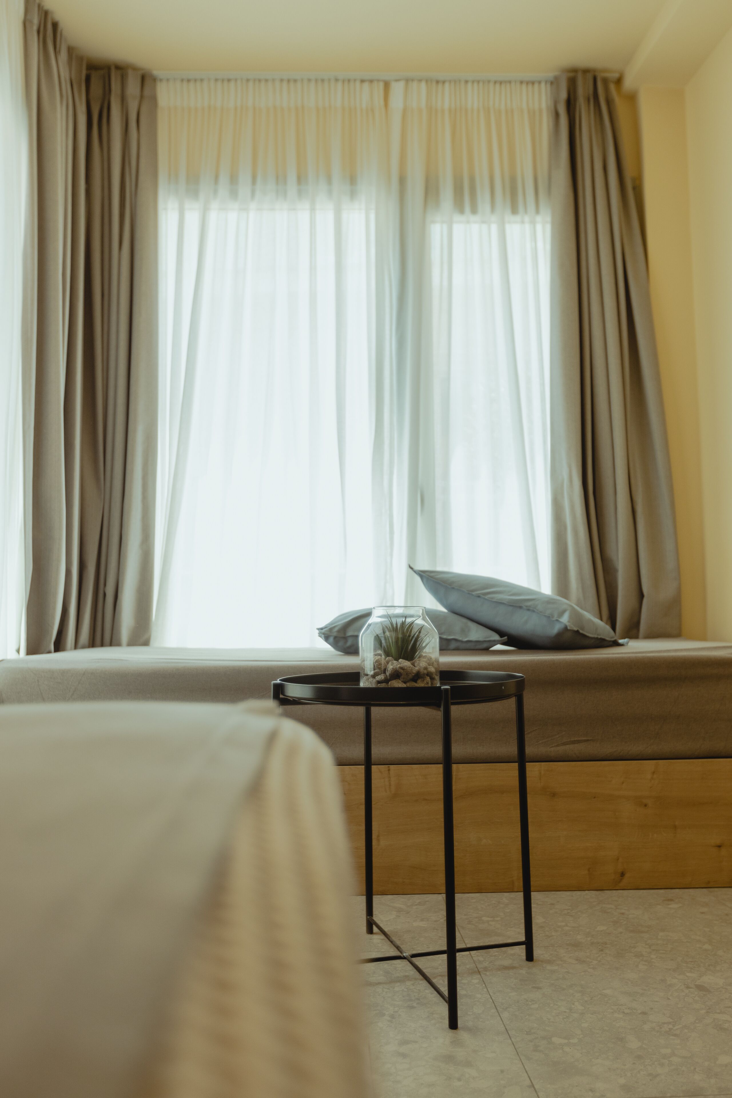 Un dormitorio con una cama, una mesita con un recipiente de vidrio y una ventana con cortinas.