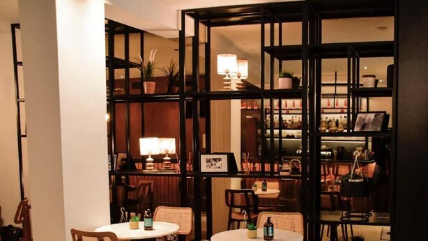 Uma área de refeições acolhedora, com mesas redondas e cadeiras de vime, um bar com estantes e plantas em vasos, e uma iluminação ambiente aconchegante.