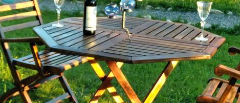 Una mesa de madera con una botella de vino y dos copas, colocada al aire libre sobre una zona cubierta de césped y sillas.