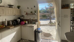 A compact kitchen with white cabinets, a sink, and a stainless steel trash bin. There are plants on the counter and a view of a garden outside. - VILLA PROVENÇALE with private pool (between Aix and Marseille) (VELAUX)