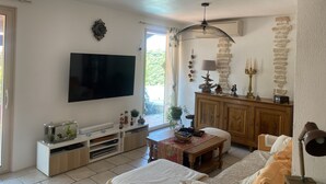 A living room with a flat-screen TV, wooden furniture, a stone wall, and a window with a view of greenery. - VILLA PROVENÇALE with private pool (between Aix and Marseille) (VELAUX)