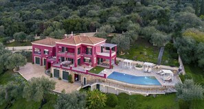Una villa rosa de varios pisos con piscina y zona de estar al aire libre, rodeada de abundante vegetación.