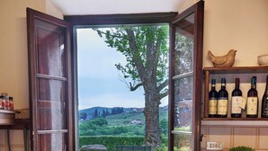 Una cocina con ventana abierta, una mesa con un bol, hogaza de pan, botella de aceite de oliva y una cesta de tomates.