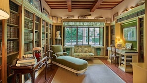 Una biblioteca con estanterías, un sillón verde, un escritorio con una silla y una ventana con vistas al verdor.