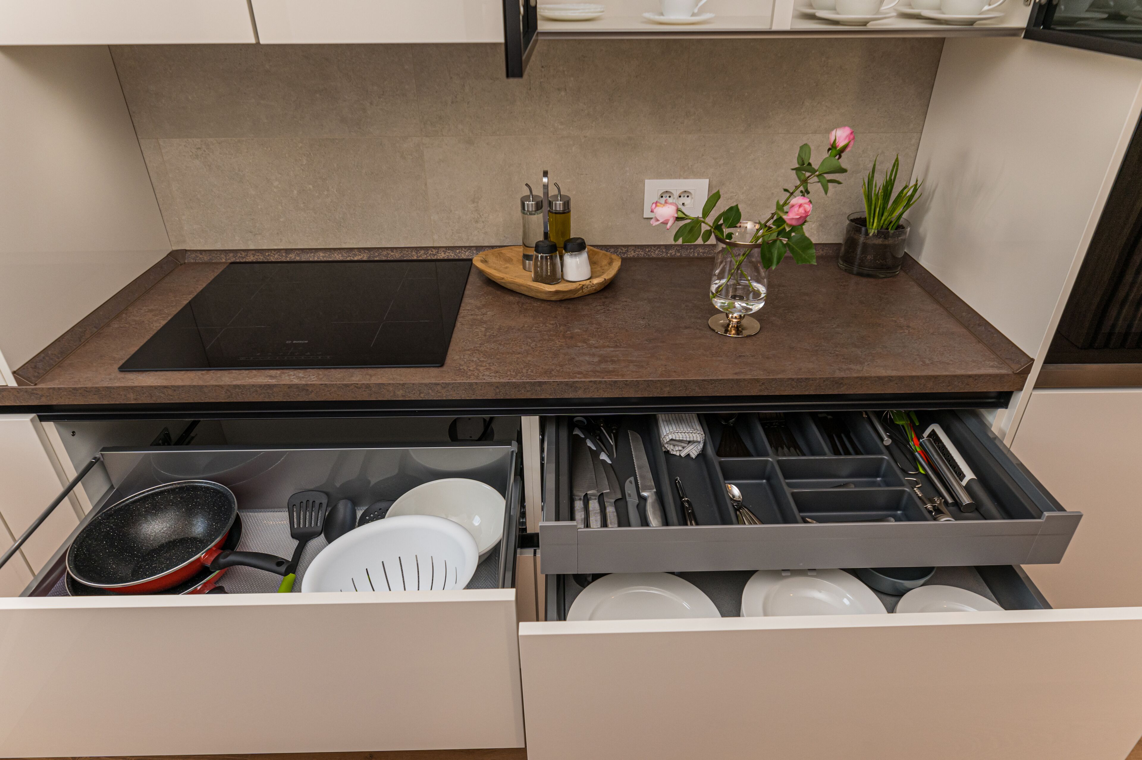 Eine Küche mit einer schwarzen Induktionskochplatte, einer Holztablett mit Salz- und Pfefferstreuern, einer Glasvase mit einer rosa Blume und einer Schublade voller Küchenutensilien.